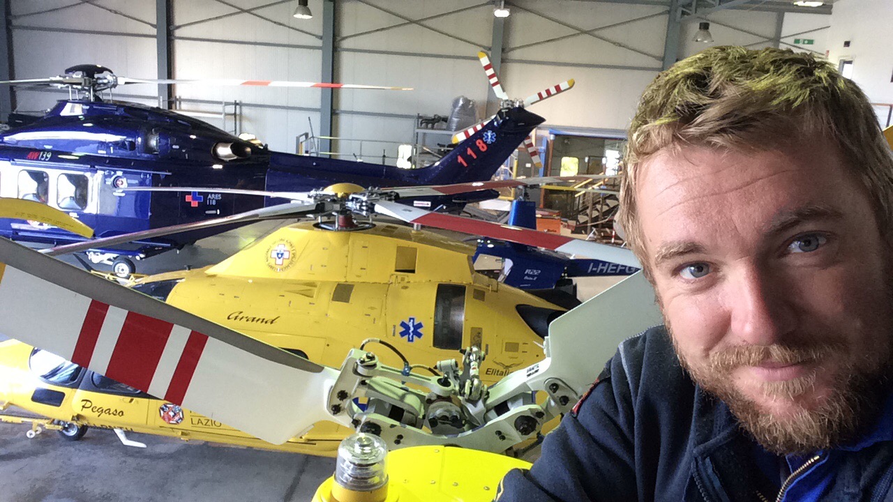 Jay Meakin takes a selfie with various helicopters in a warehouse